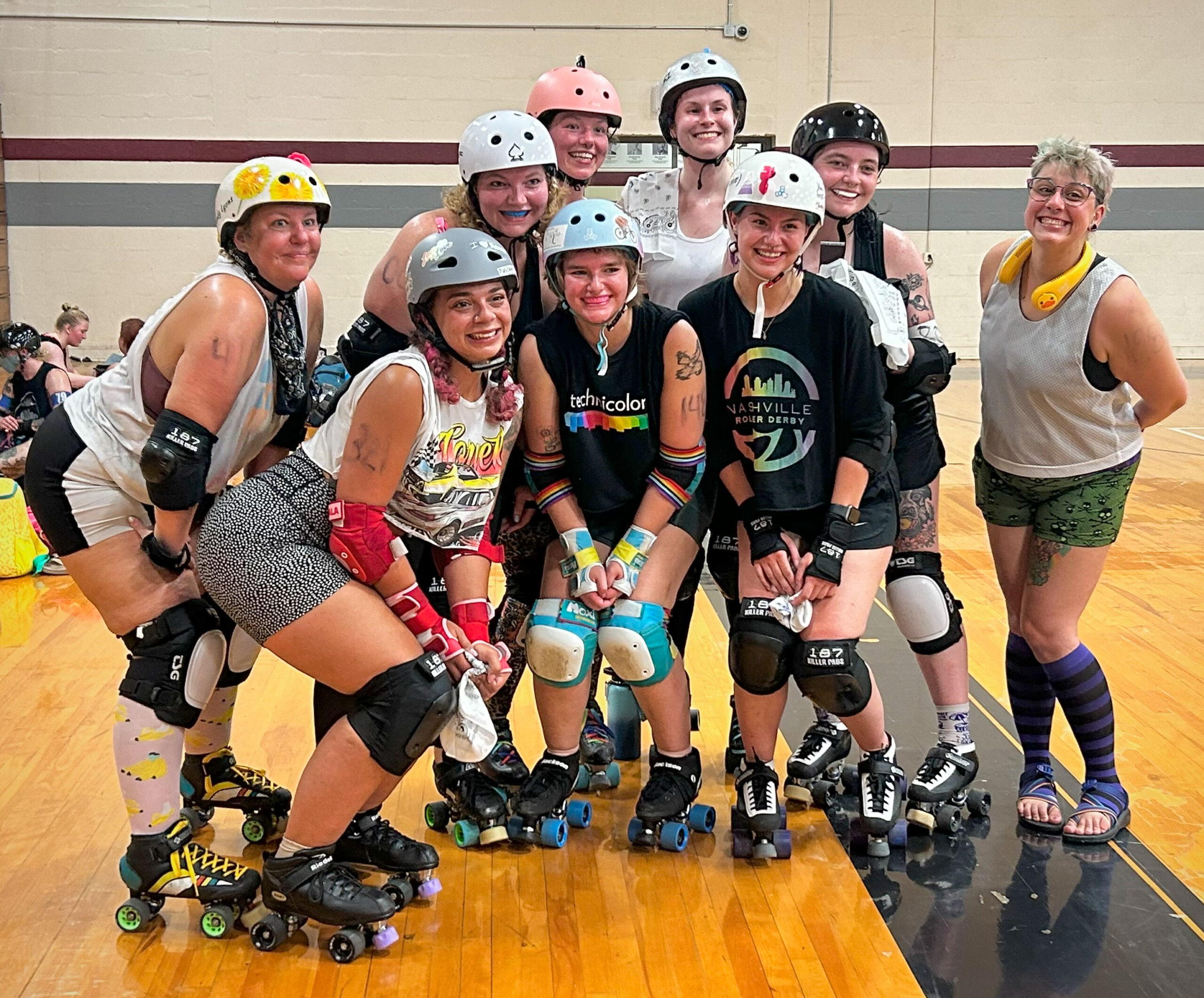 A group of nine people are posed together on a roller derby track. Eight are wearing roller skates, helmets, knee pads, elbow pads, and wrist guards, with smiles on their faces. One person is standing to the far right, not wearing skates, in casual athletic attire, purple and black striped knee-high socks, and sandals. Most participants wear a mix of colorful, casual athletic clothing, including shirts and shorts, with various patterns and designs. The group appears happy and energetic, suggesting a fun or celebratory moment during a Nashville Roller Derby bootcamp session.